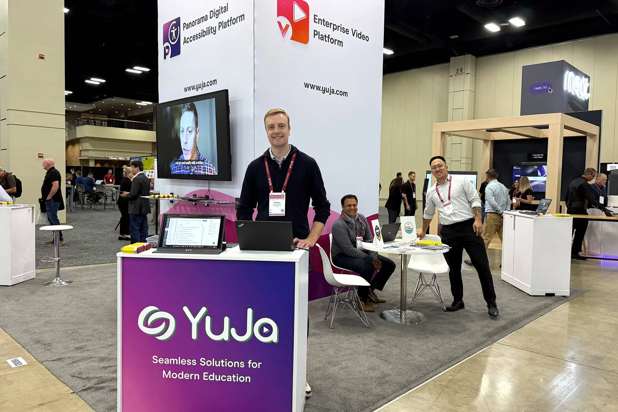 People congregated around YuJa's booth displaying a laptop and a white sign, actively participating in a discussion.