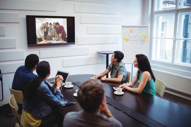 People in a conference room watching a video monitor hanging on the wall.