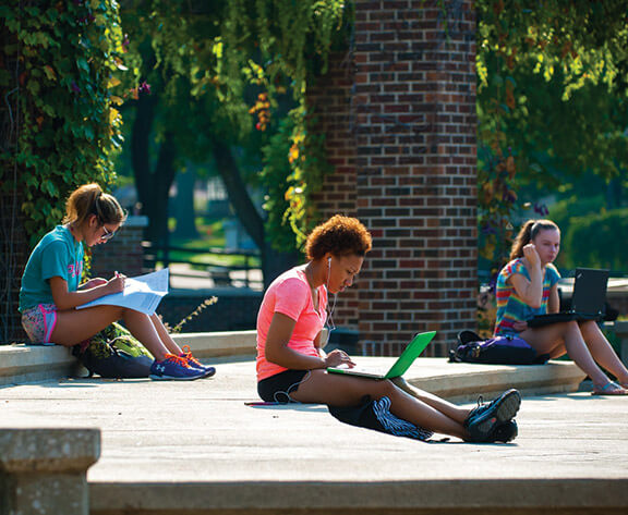 Three students from Indiana State University sitting on a bench, engrossed in their laptops.