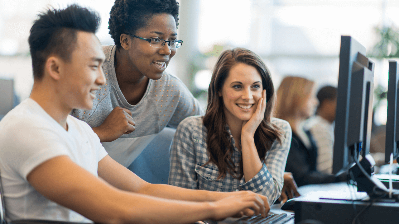 Three young people smiling while working on computers in a modern office.