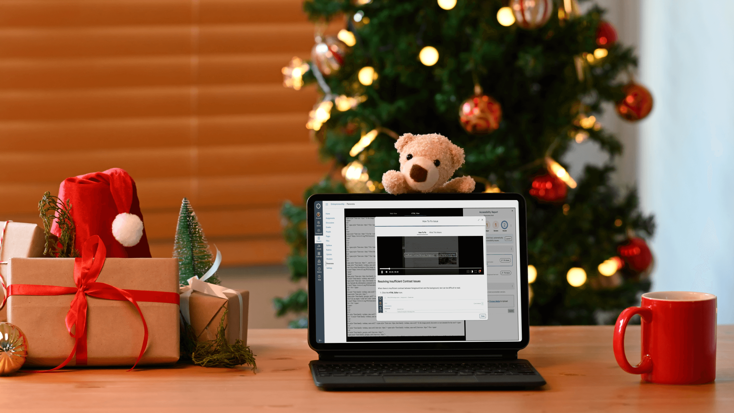 A laptop featuring an LMS-specific How to Fix video sits on a desk in front of a holiday tree, with wrapped presents on one side and a mug on the other. A teddy bear hangs off the laptop.