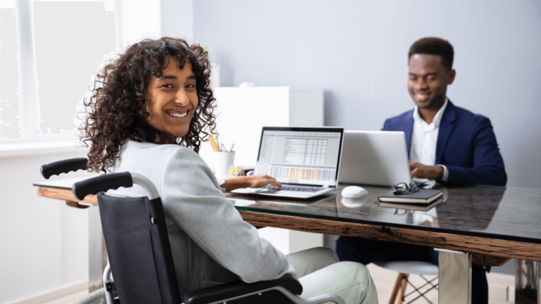A young person in a wheelchair looks at a laptop. Across the table is another student looking at a laptop.