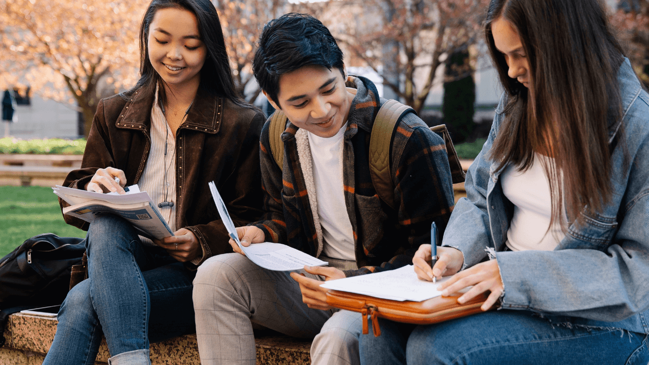 Three students sitting on a bench outdoors, focused on writing notes in their notebooks.