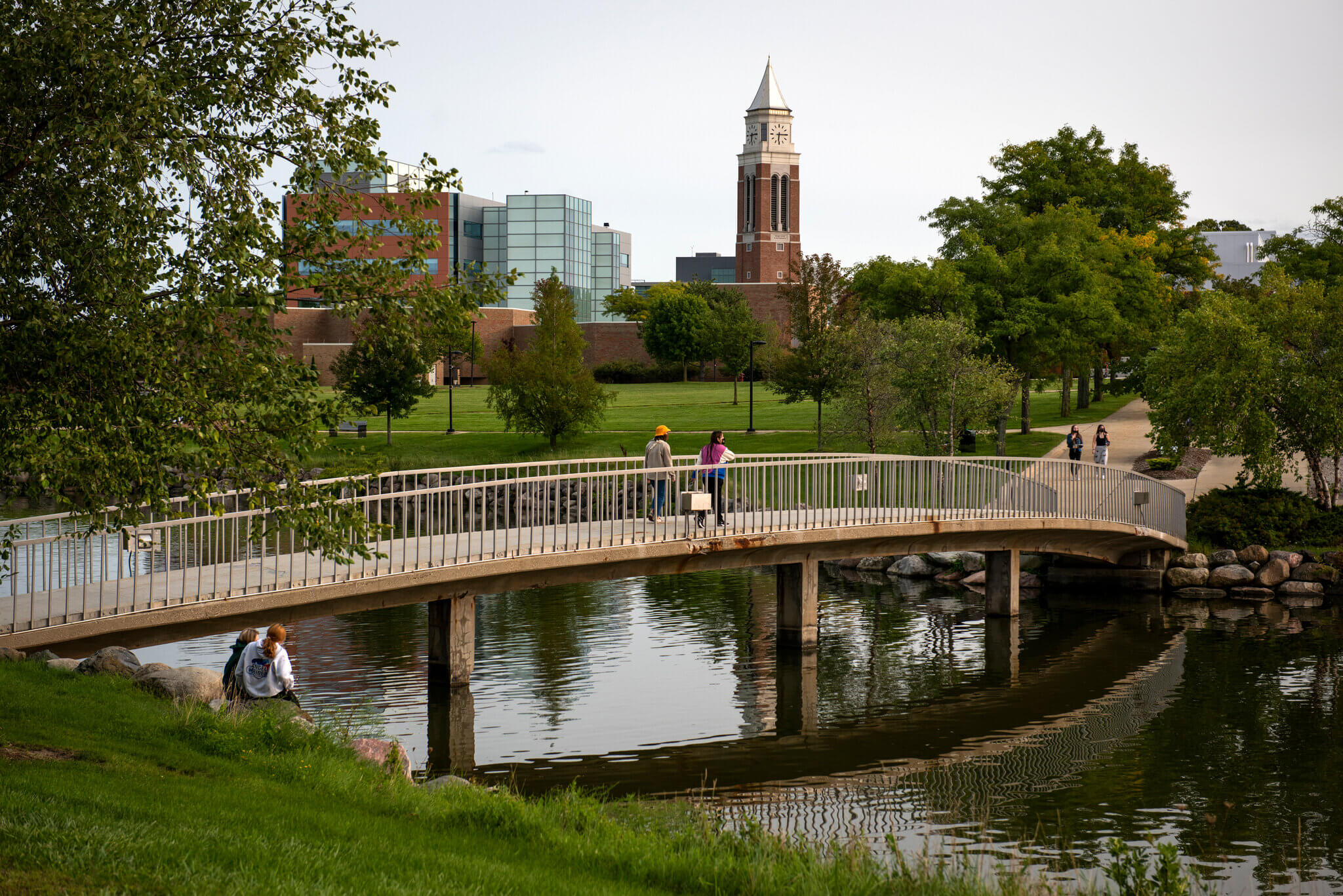 People walking across a bridge over a serene pond with Oakland University in the background.