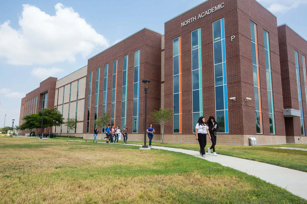 Four South Texas College graduates raising their diplomas while wearing their graduation hats and gowns in front of a campus building.