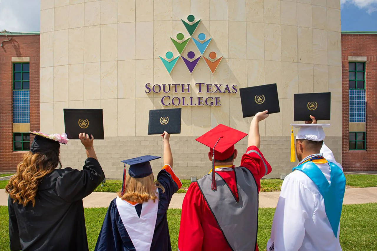 Four South Texas College graduates raising their diplomas while wearing their graduation hats and gowns in front of a campus building.