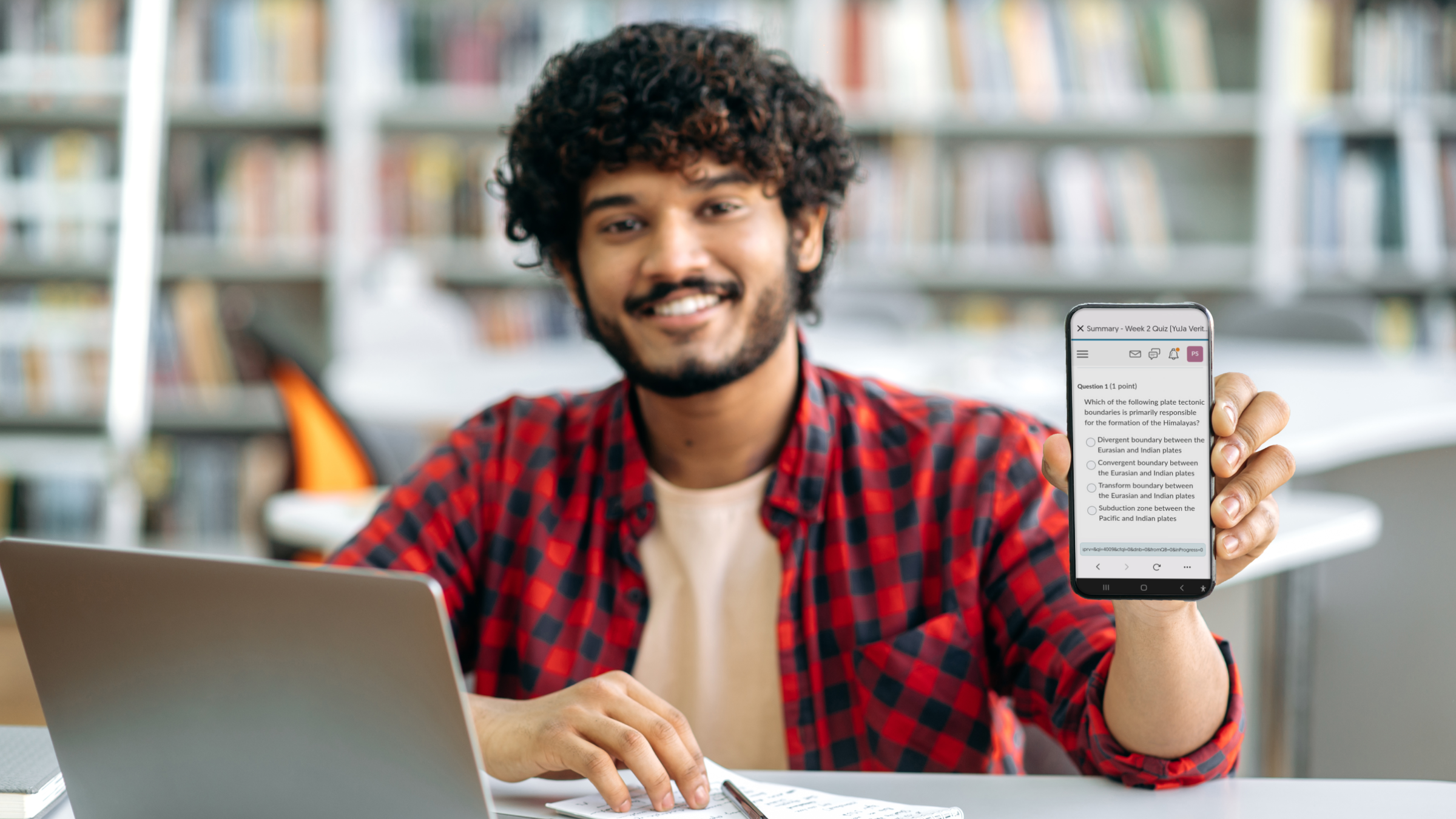 A college student sitting at his desk, in front of a laptop, and holding out his phone, which features the Verity Mobile Lockdown App support for D2L Brightspace.
