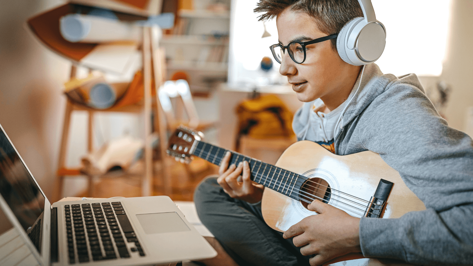 Student wearing headphones and holding a guitar looks at a laptop.