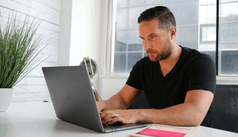 A man looks at a laptop with a clean white bacdrop and windows behind him.
