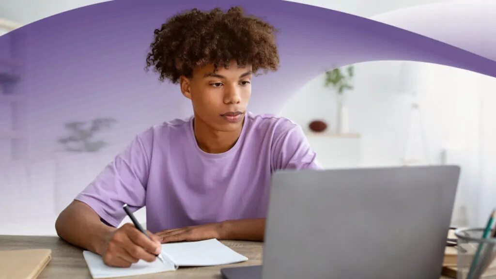 A young man sitting at a desk with a pen and a notebook, looking at his laptop.