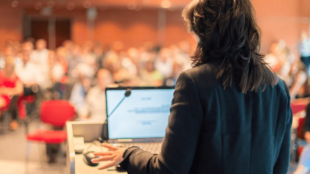 This image shows the back of a lecturer at a podium. You can see their laptop screen and a microphone. The background is a full lecture hall.