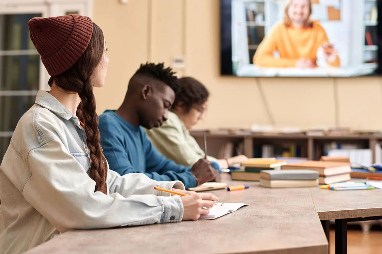 Three students sitting in a lecture watching a video of a man with a female student looking at the screen.