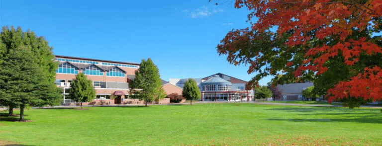 A view of a green field, with NHTI Concord’s Community College buildings in the distance and surrounding trees.