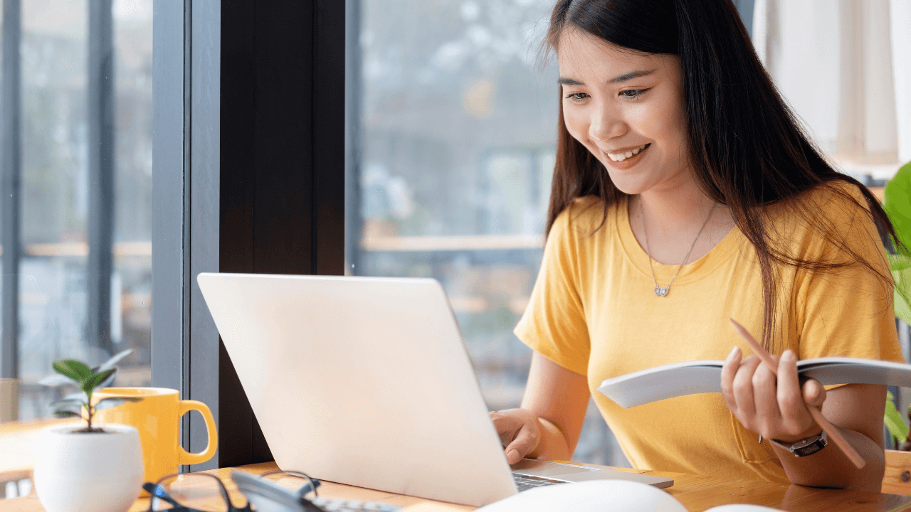 A young woman looks at a book and her computer.