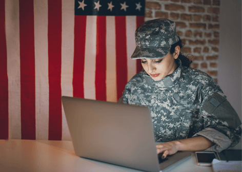 Woman in military fatigues sits at a laptop with an American flag in the background.