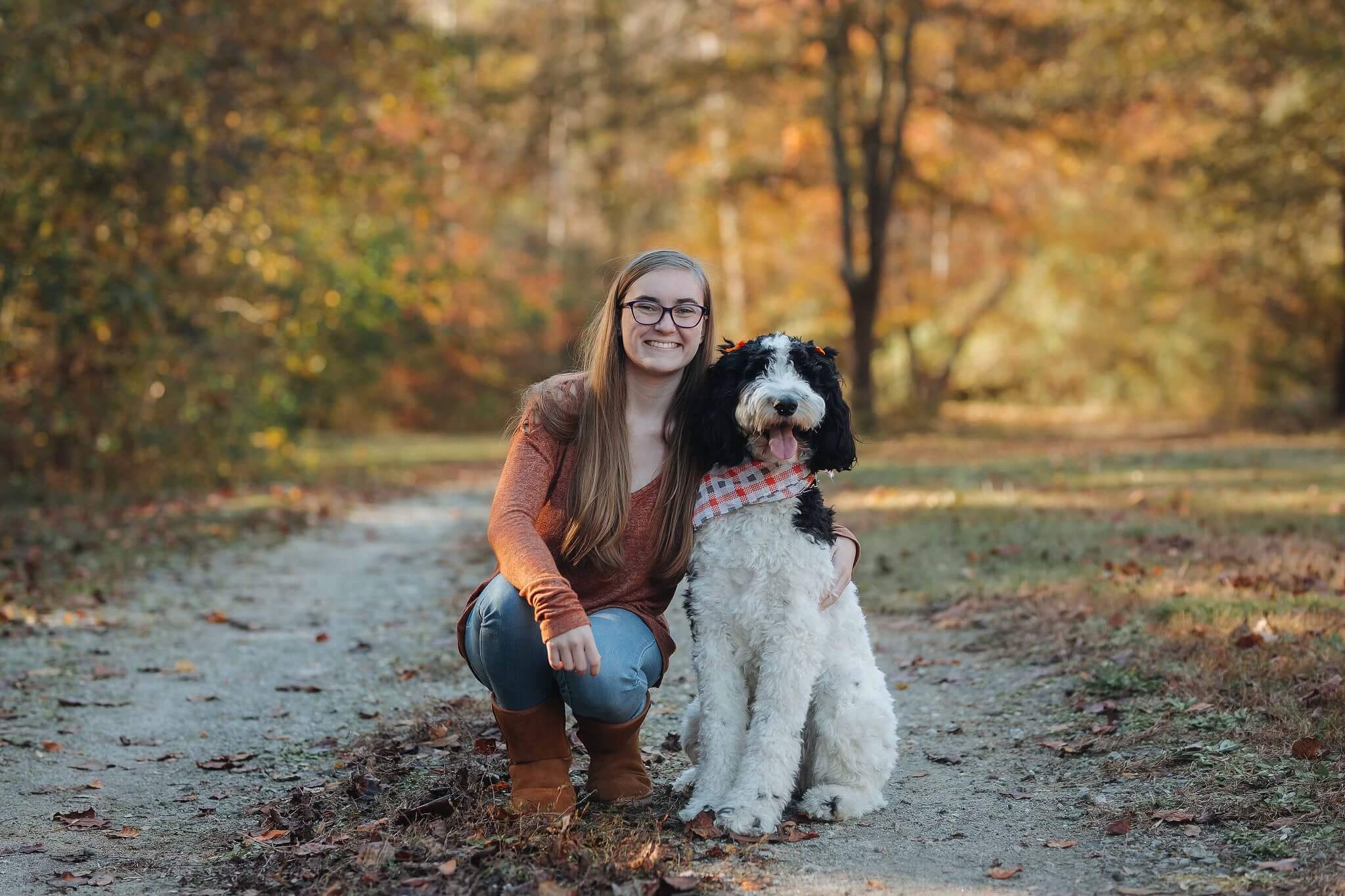 College student poses with her dog on a trail with fall colored leaves on trees in the background.