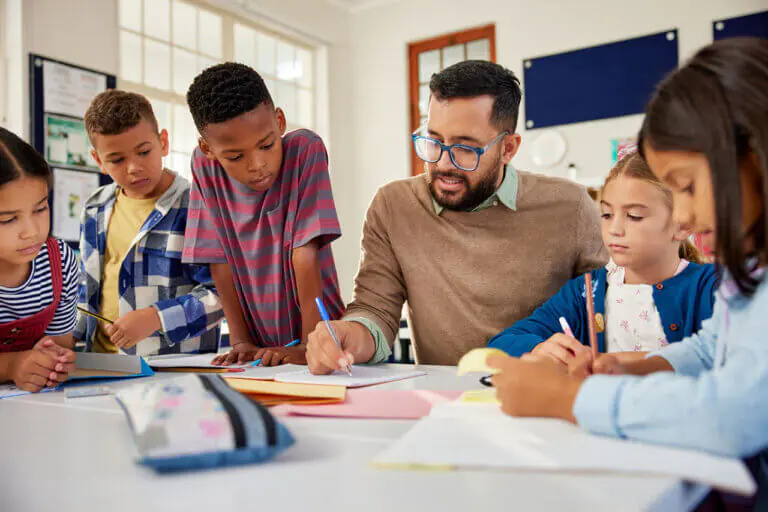 “An elementary school classroom with 5 young children sitting around an adult male teacher explaining a worksheet.