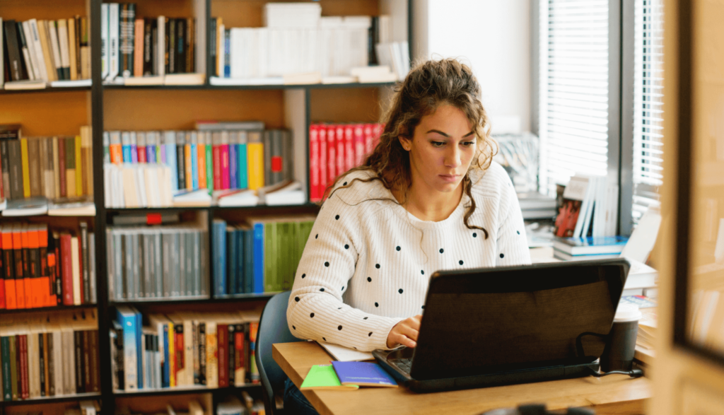 Student sits at a laptop with a bookshelf of books in the background.