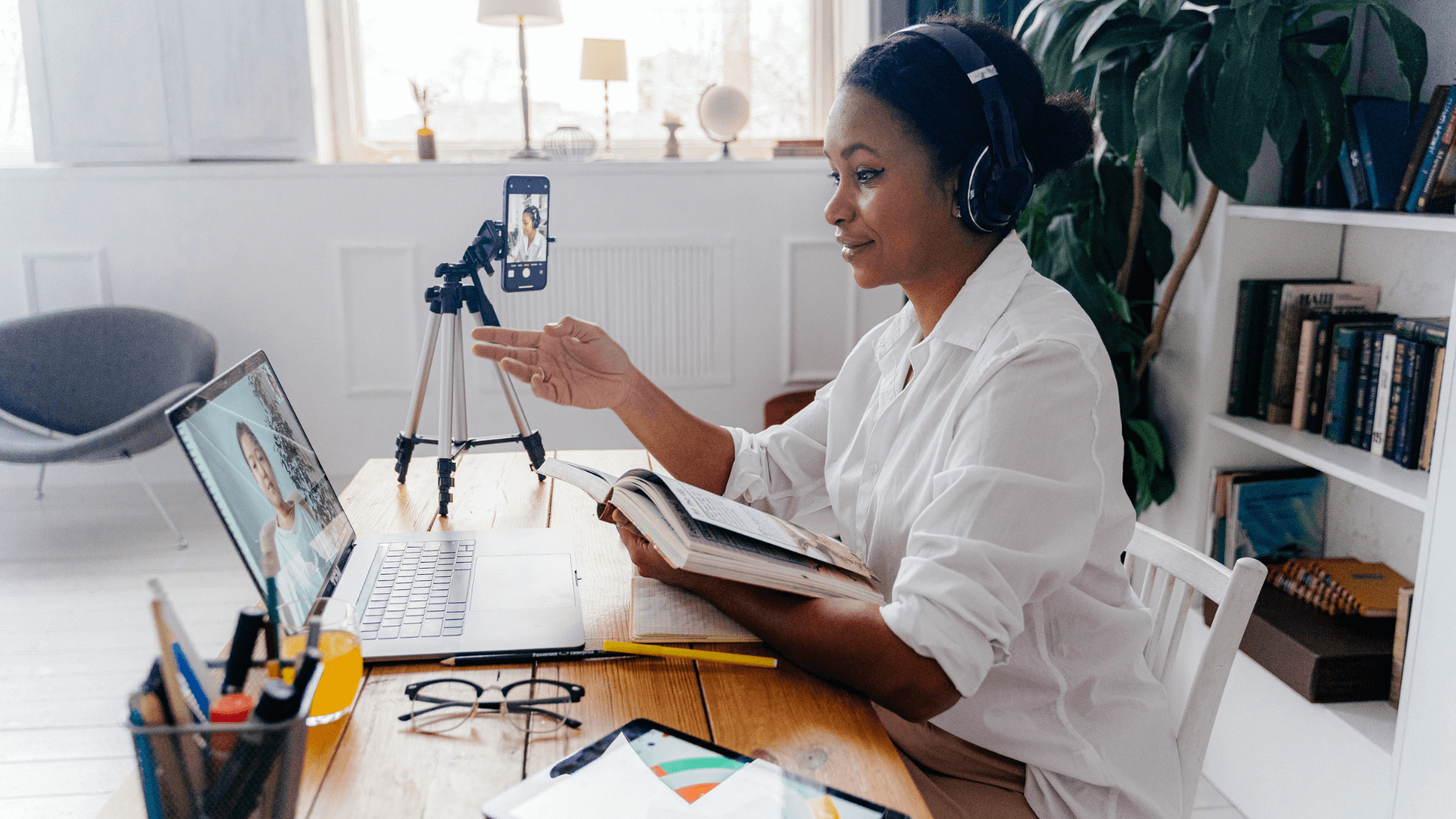 Woman wearing headphones presents in front of a laptop. A cellphone is on a tripod to record her as well.
