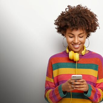 A woman wearing headphones and a vibrant sweater is focused in her phone.