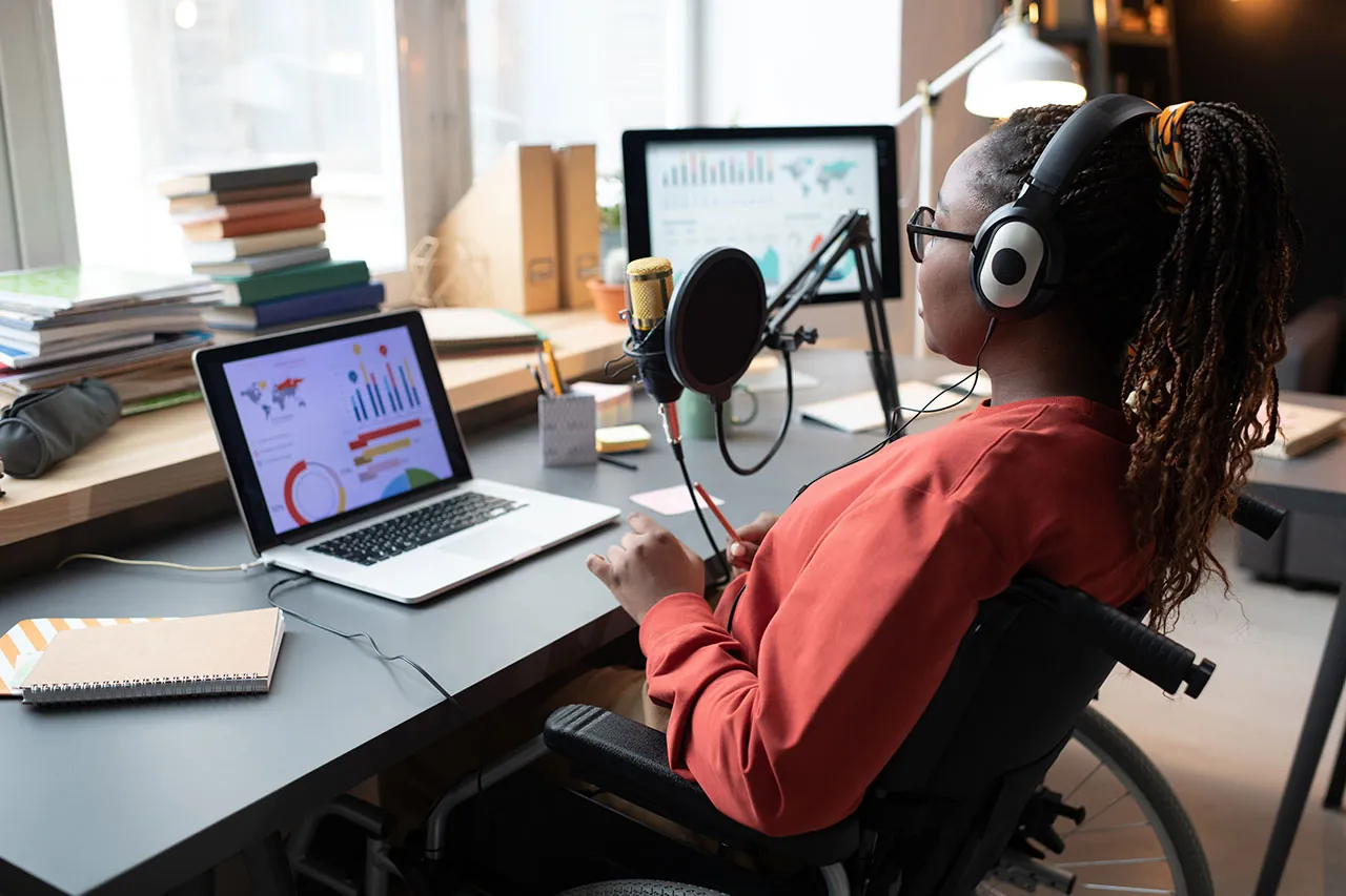 A woman with headphones sitting in a wheelchair in front of a laptop with graphs, speaking into a microphone.
