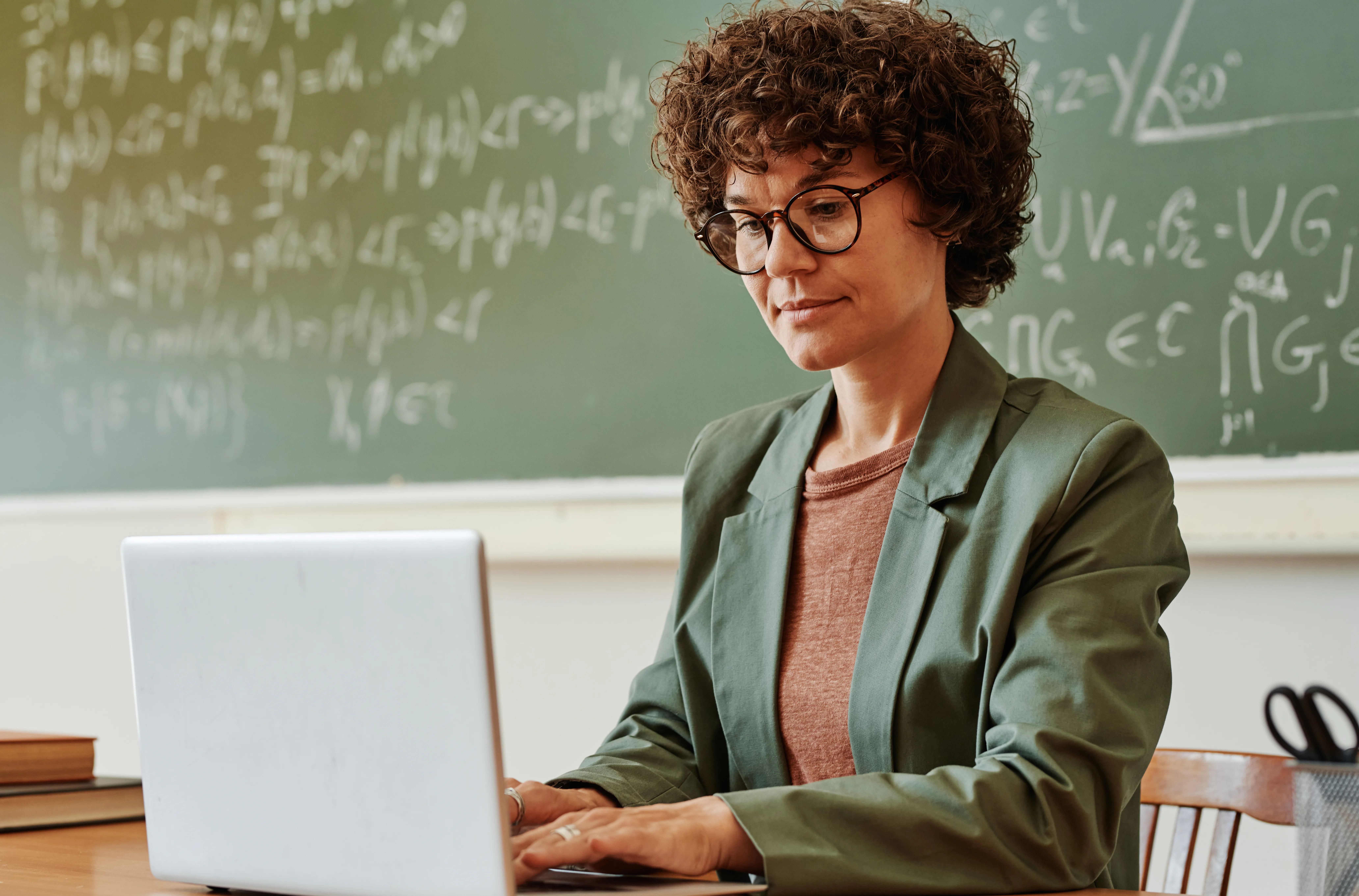 Young confident teacher typing on laptop keypad while sitting in front of a blackboard with formulas and equations.